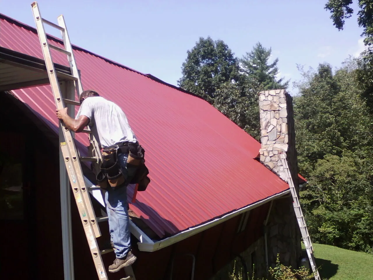 Roofer on a ladder beside a red metal roof and stone chimney — Soffit & Fascia Repair by Roofing Brothers in Marne